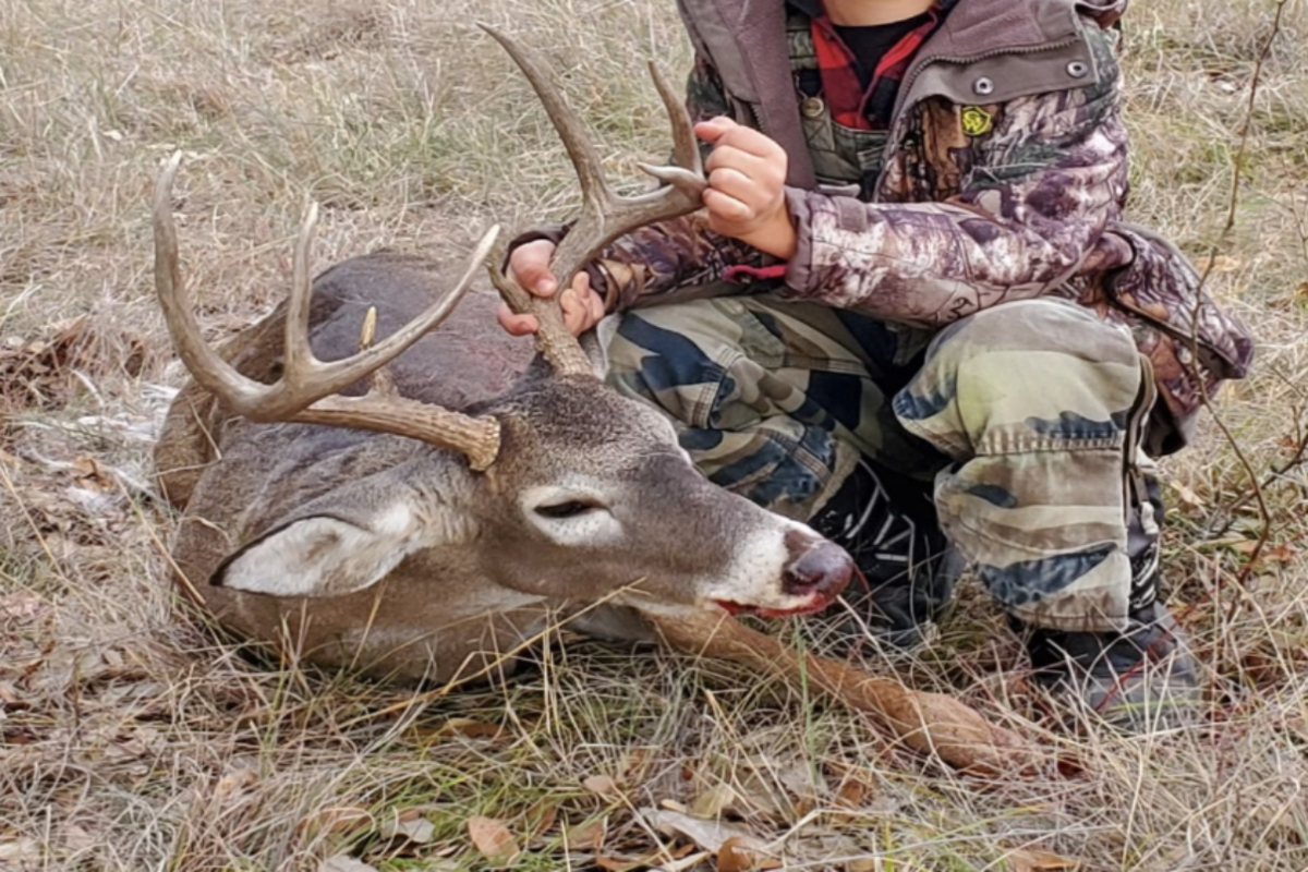 A person in camouflage hunting clothing kneels beside a large buck deer lying on dry grass and leaves outdoors.